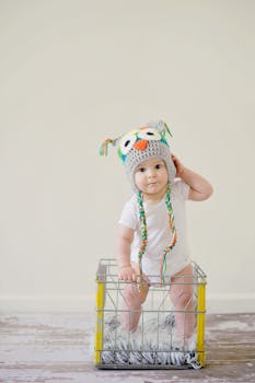 A cute baby wearing an owl hat standing in a basket, smiling and playful.